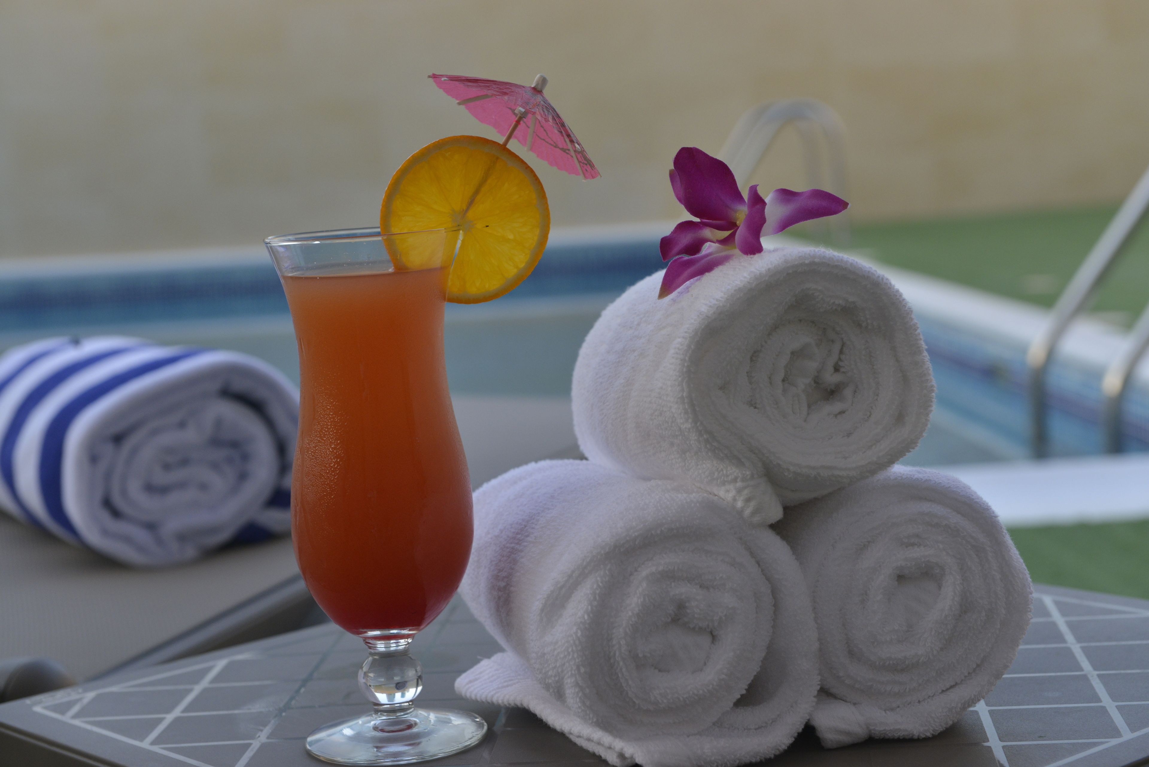 A glass of orange-colored drink with a slice of orange and a pink umbrella, next to rolled white towels and a striped towel on a table by a pool.