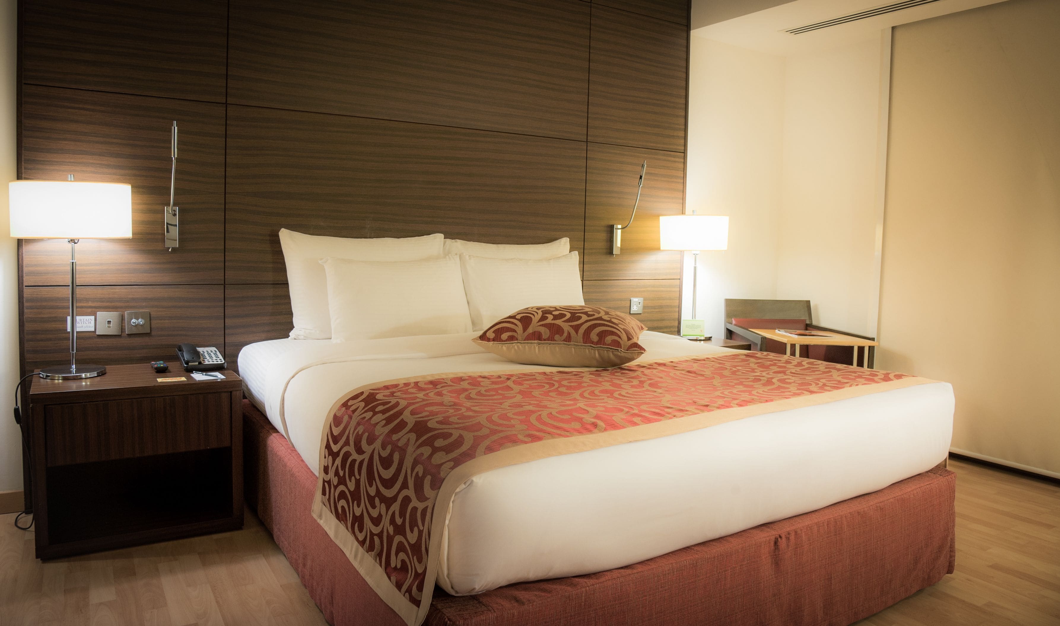 A hotel room with a large bed, two bedside lamps, a nightstand with a phone, and a desk with a computer.