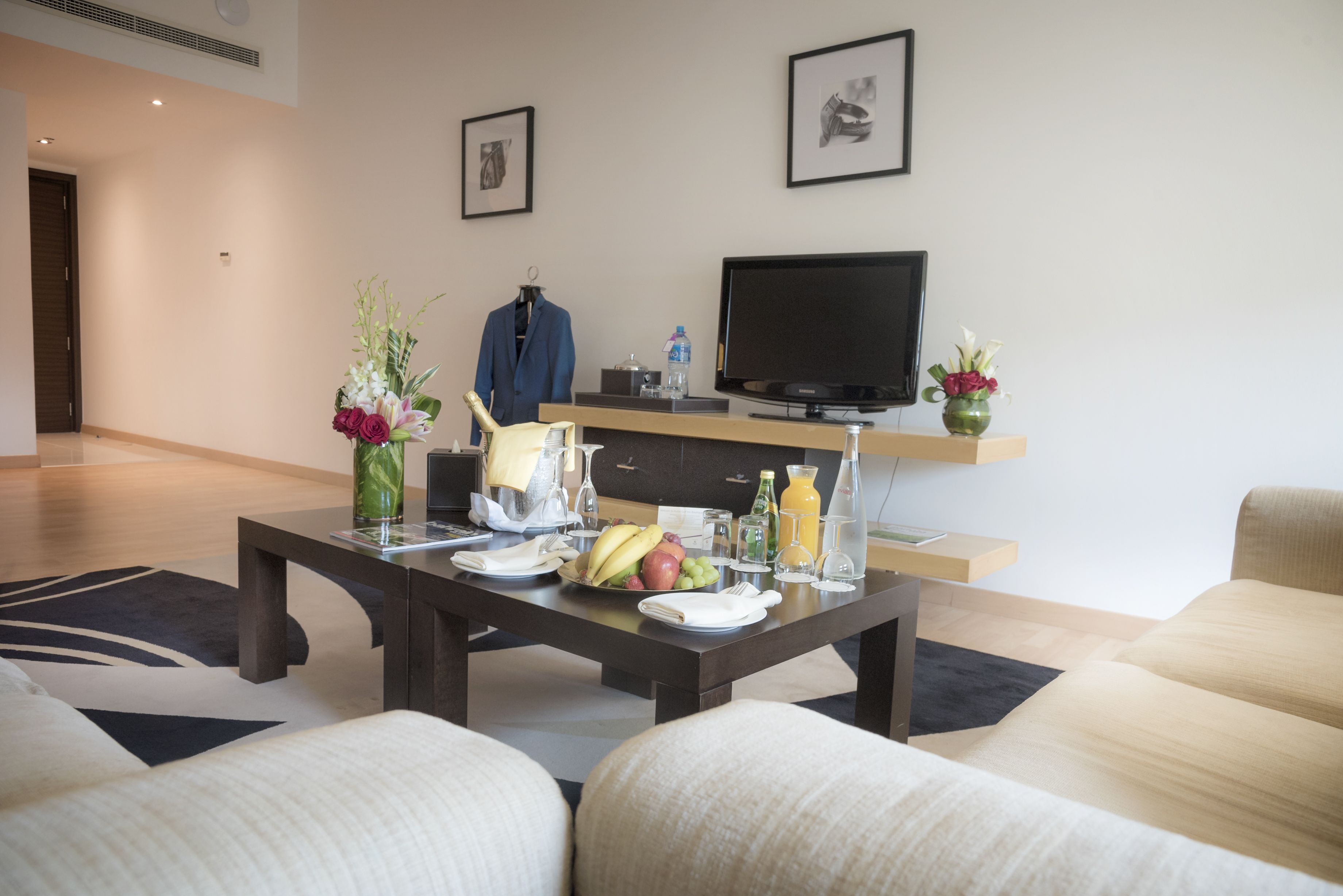 Una sala de estar moderna con un sofá, una mesa de centro y un televisor de pantalla plana. Se observa un perchero con una chaqueta azul, un jarrón con flores y un frutero sobre la mesa de centro.