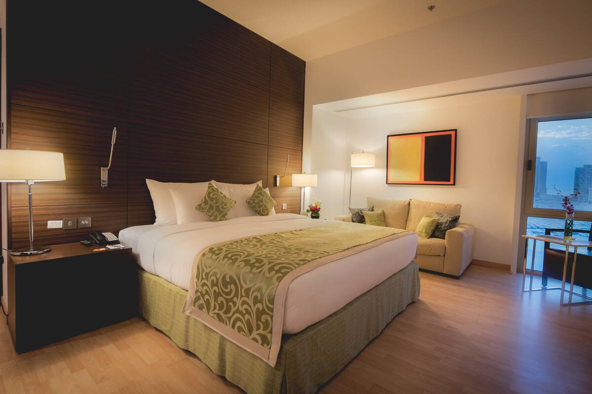 A hotel room with a large bed, bedside lamps, a sofa, a small table, and a view of the city through a large window.