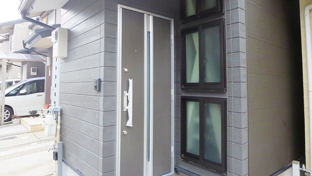 Une maison moderne avec une façade grise, une porte d’entrée dotée d’une poignée et trois fenêtres aux montures sombres.