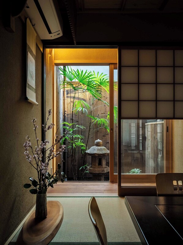 Un intérieur japonais traditionnel avec un sol recouvert de tatamis, une table basse, une chaise, un vase de fleurs et une vue sur la verdure à l’extérieur.