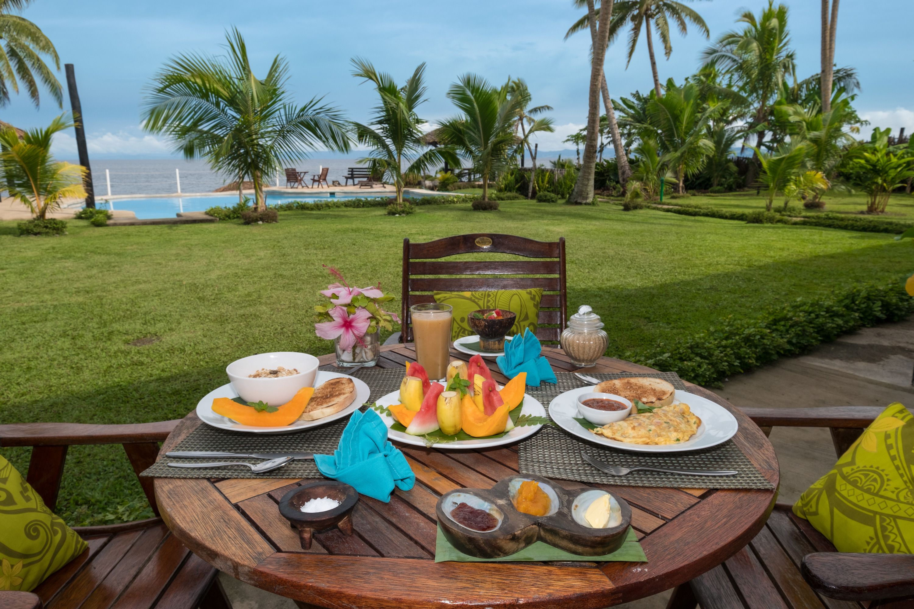 A wooden table set with breakfast items, including fruits, bread, and beverages, overlooking a pool and palm trees.