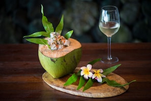 A wooden table with a coconut shell containing a dish, a glass of white wine, and a flower. - Paradise Taveuni (Taveuni Island West)
