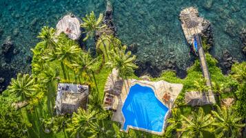 Relax by the clear blue pool surrounded by lush greenery and palm trees, with a shaded area under a thatched-roof structure.