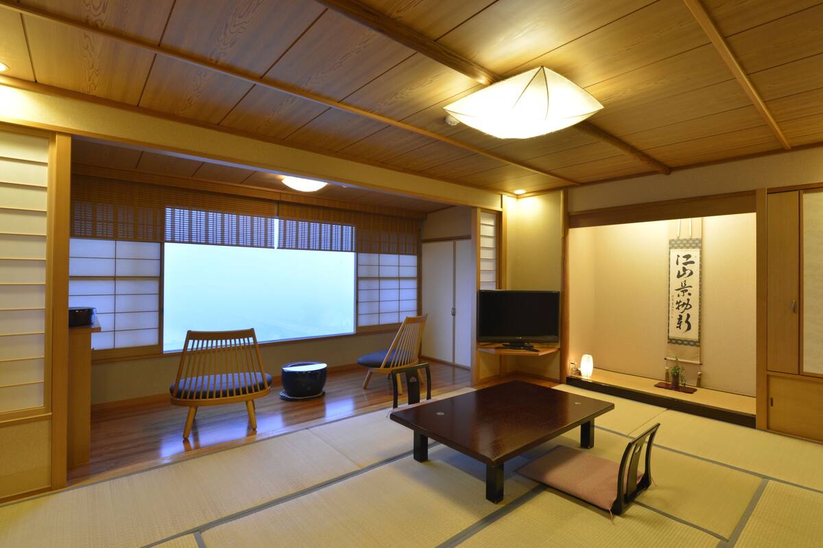 Een traditionele Japanse kamer met tatami vloerbedekking, een lage tafel, twee stoelen, een televisie en schuifdeuren.