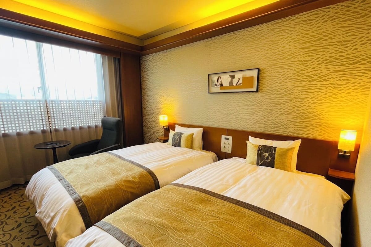 Een hotelkamer met twee bedden, een klein tafeltje en een stoel. Er is een raam met jaloezieën, een gestructureerde muur en een ingelijste foto aan de muur.