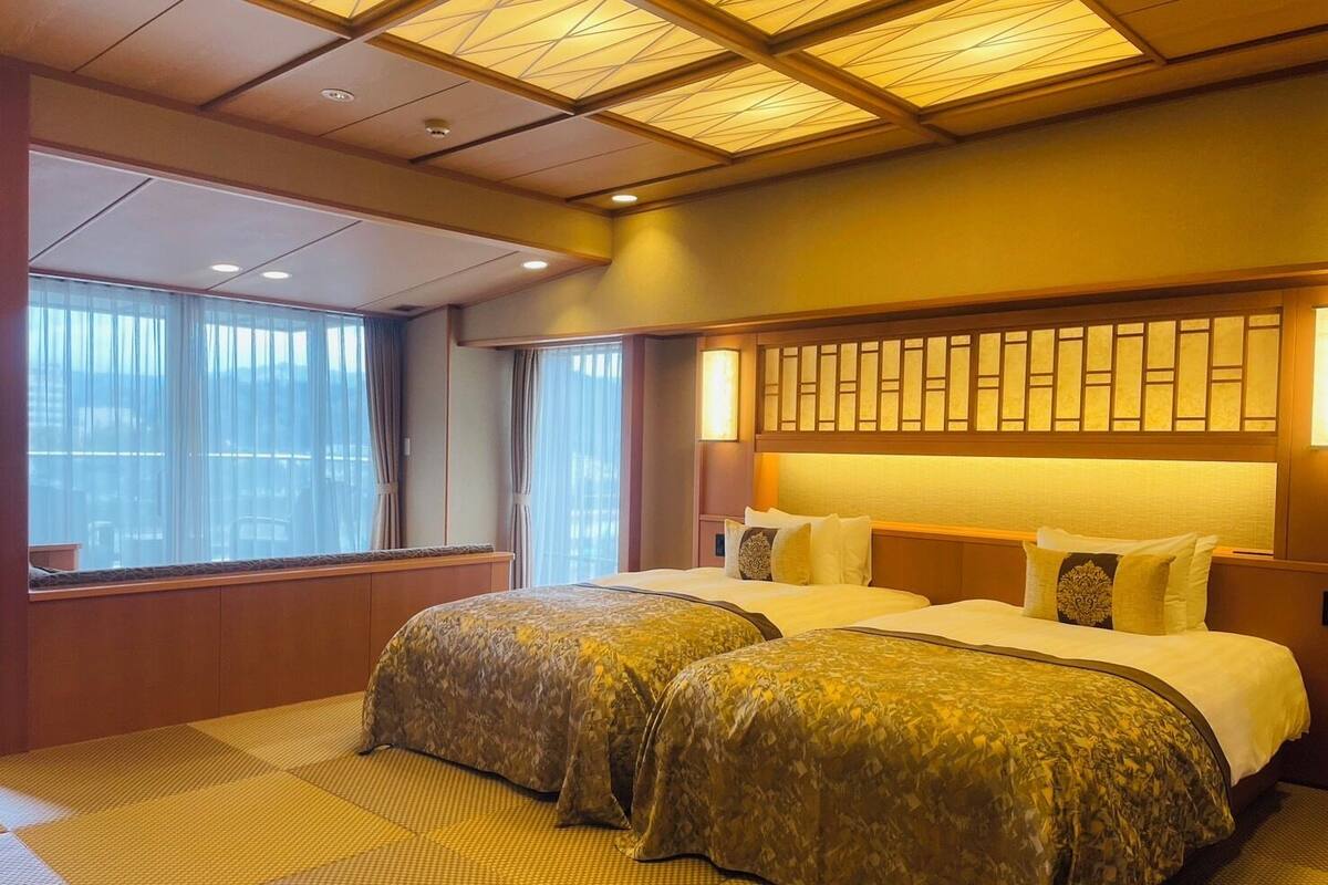 Een hotelkamer met twee bedden, een groot raam met gordijnen, een plafond met een geometrisch patroon en een wand met een decoratief paneel.