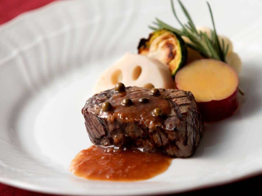 A plate with a steak topped with mushrooms and capers, accompanied by roasted vegetables and a slice of apple.