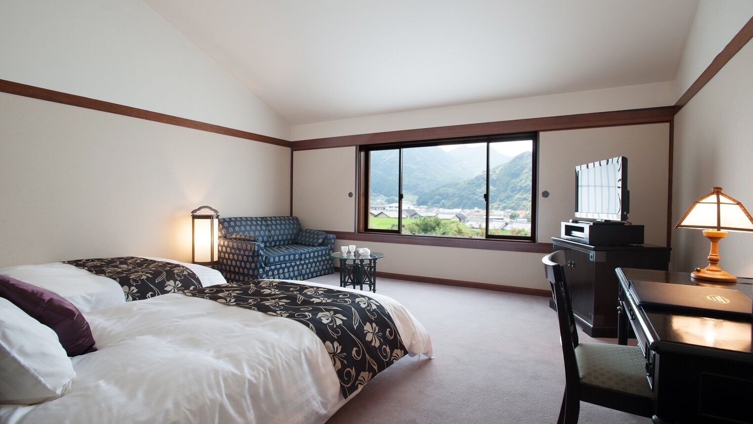A hotel room with a bed, a desk, a chair, a small table, a lamp, and a large window with a view.