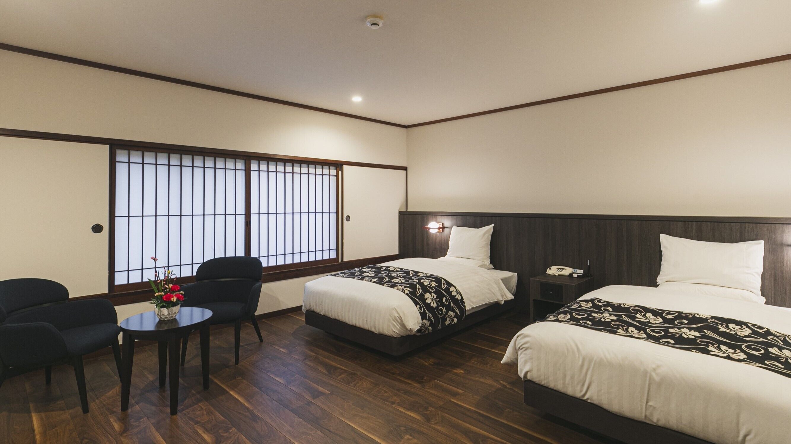 A hotel room with two beds, a small table with a vase, a chair, and a window with a view.