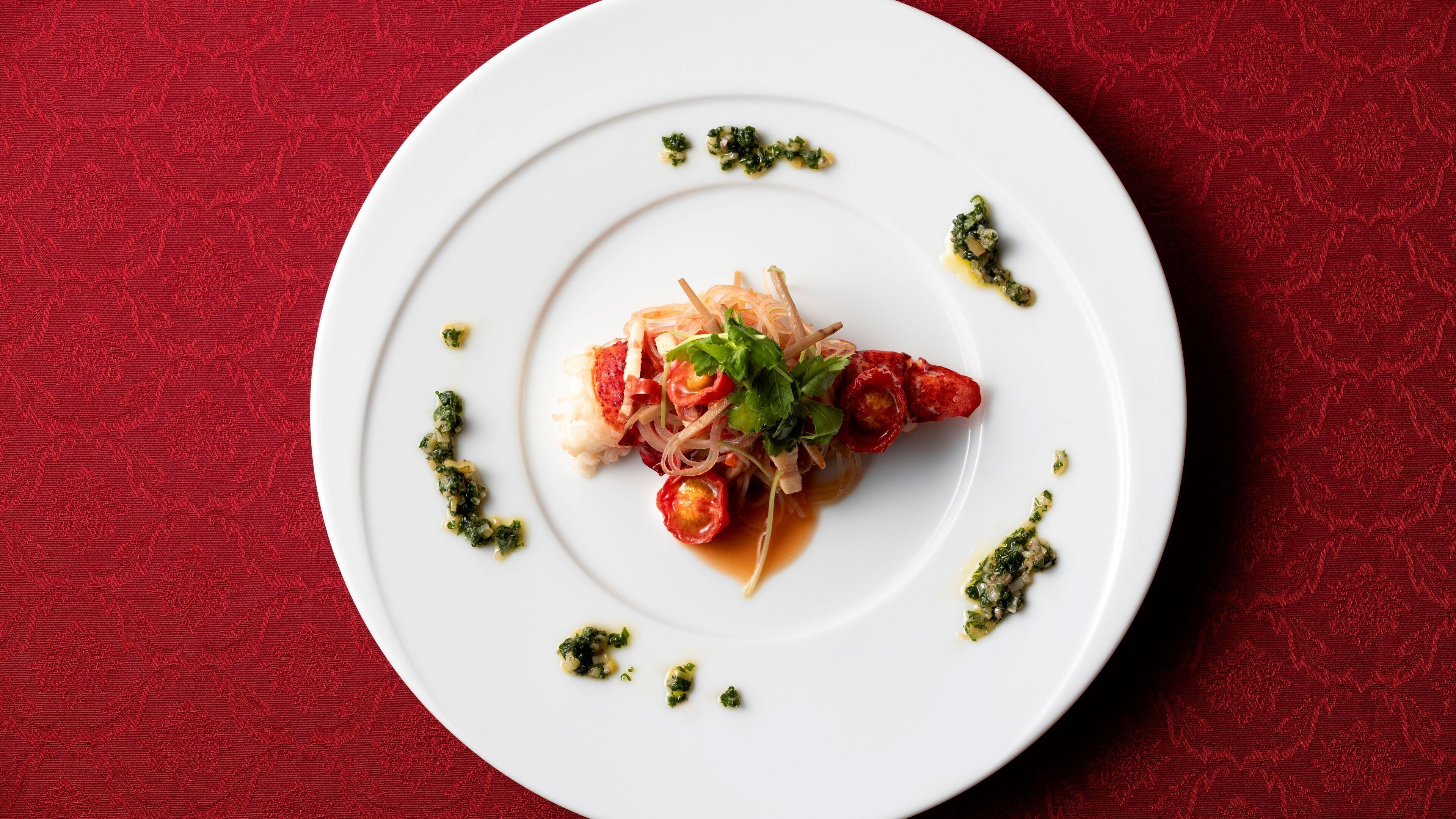 Un plato blanco con un guiso de comida, que incluye pescado, verduras y hierbas, sobre un fondo rojo con textura.
