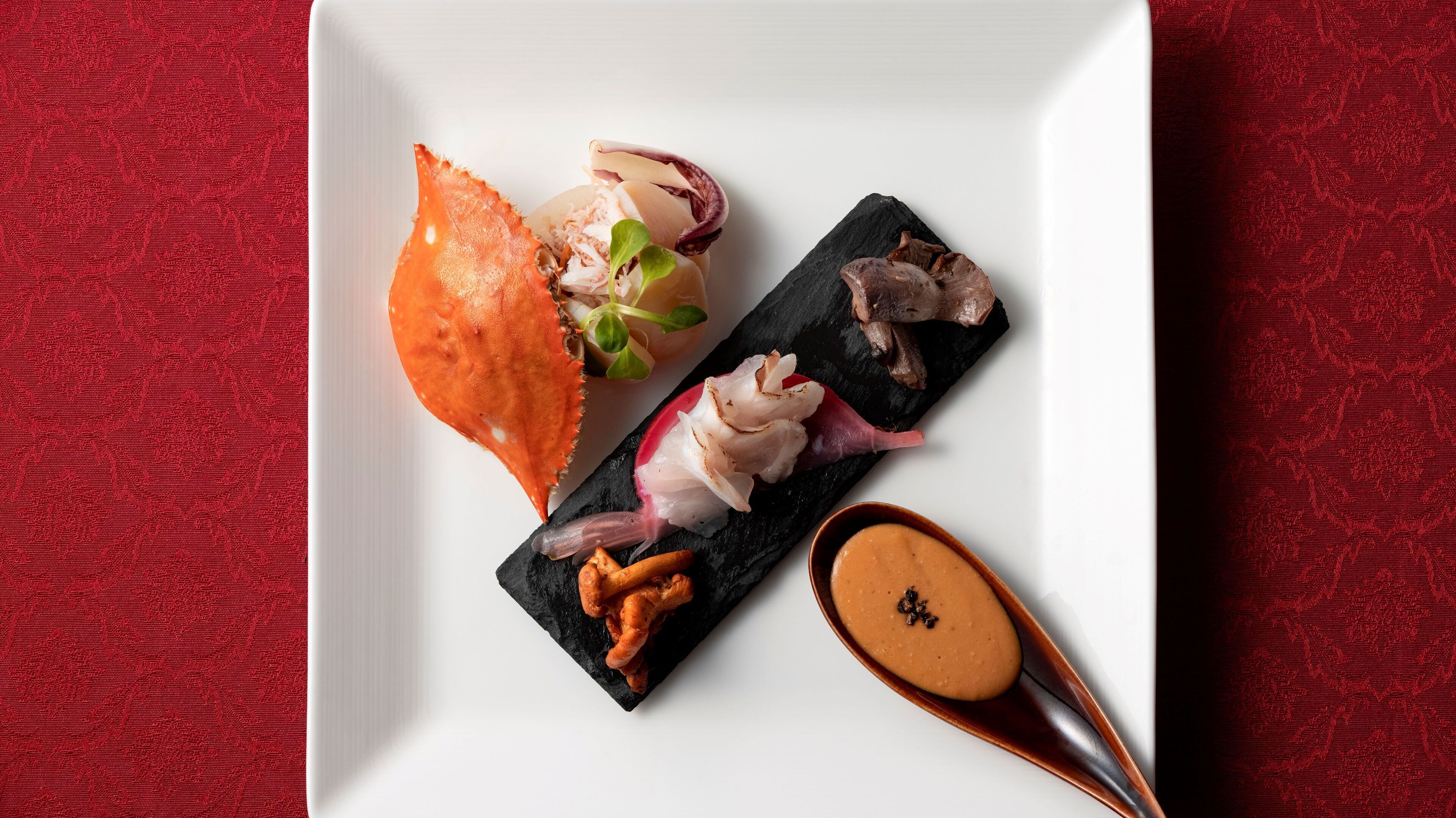 Un plato blanco cuadrado con diversos alimentos, incluyendo una pata de cangrejo, carne en rebanadas y un pequeño recipiente con salsa, sobre una superficie roja con textura.