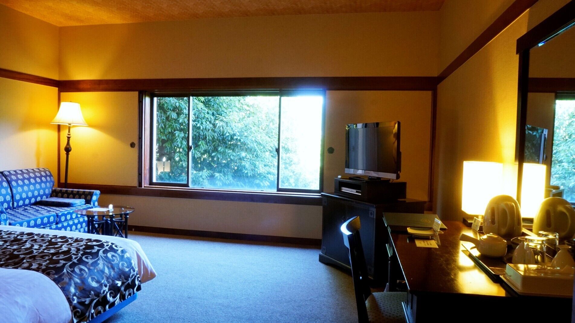 A hotel room with a bed, a desk, a television, and a window with a view of trees.