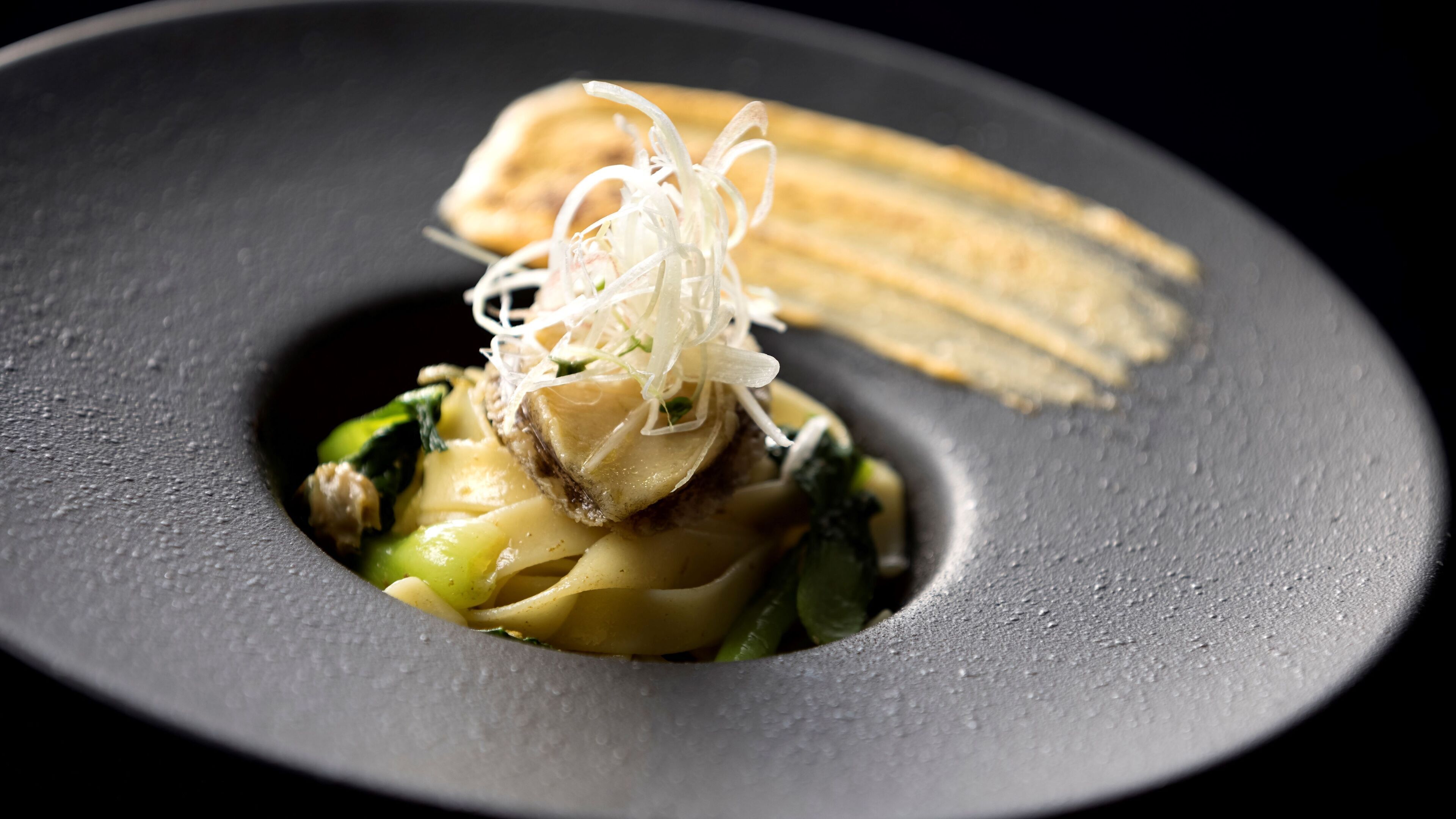 A dish with noodles, vegetables, and a piece of meat, served on a dark plate.