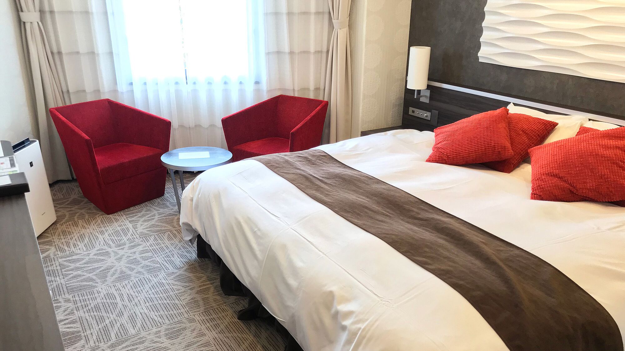 Une chambre d’hôtel avec un lit, deux chaises rouges, une petite table ronde et une fenêtre avec des rideaux.
