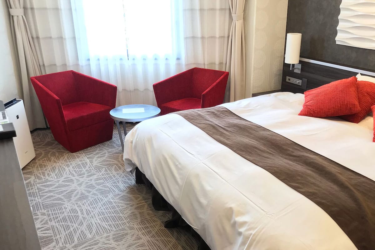 A hotel room with a bed, two red chairs, a small round table, and a window with curtains.