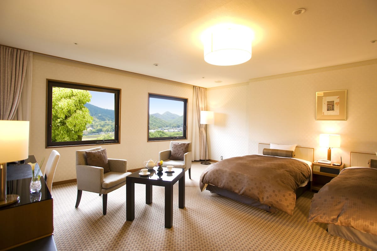 Habitación de hotel con dos camas, una zona de estar con mesa y sillas, y ventanas amplias con vistas a zonas verdes y montañas.