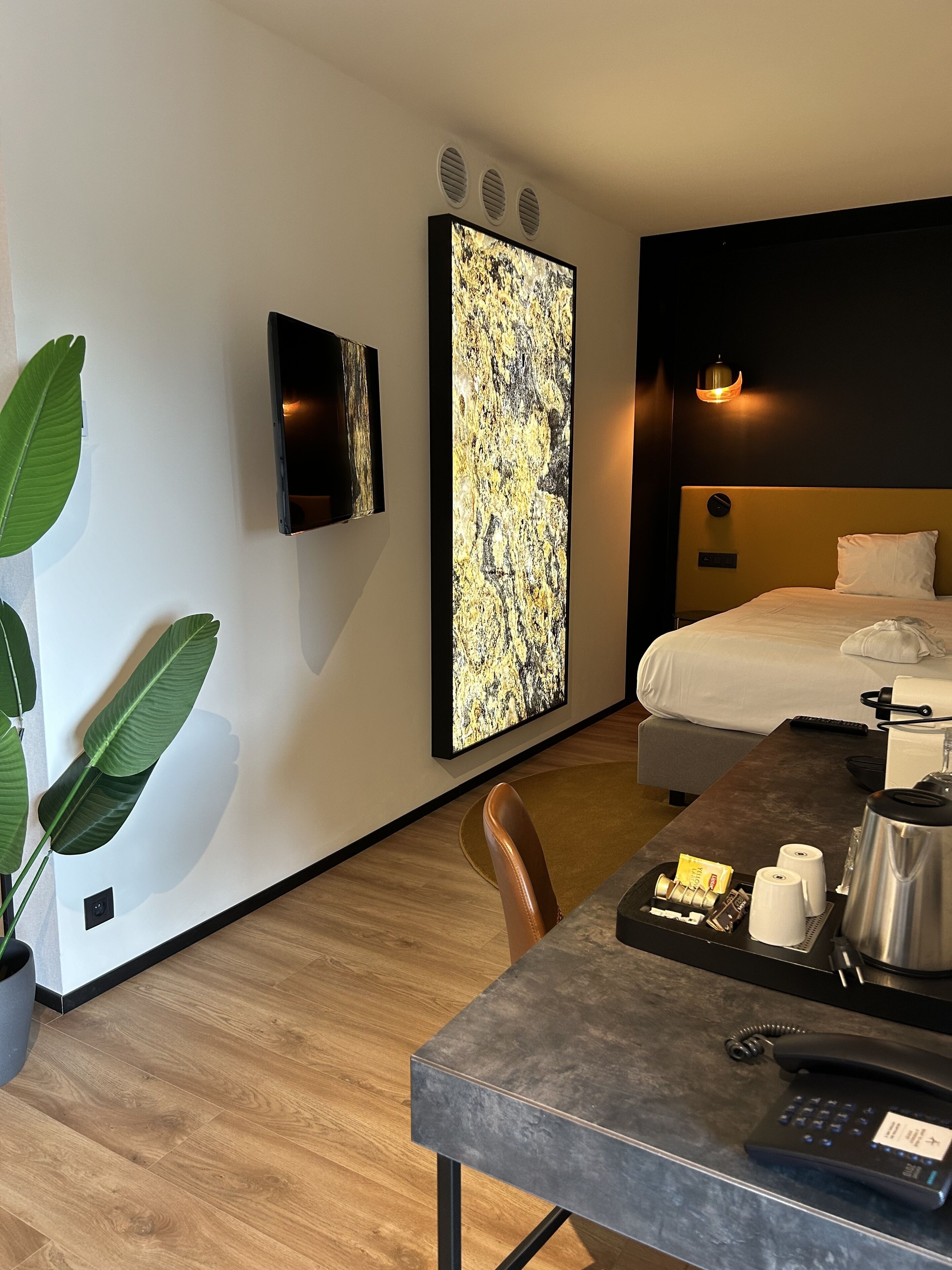 Habitación de hotel con cama, escritorio con teléfono, cafetera y un cuadro abstracto grande en la pared.