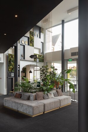 A modern lobby with a grey sectional sofa, indoor plants, and a sign indicating a bar and restaurant. - Hotel Ariane (Ypres)