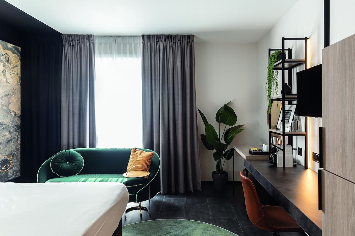 A modern hotel room with a bed, a green sofa, a desk with a computer, a plant, and a TV.