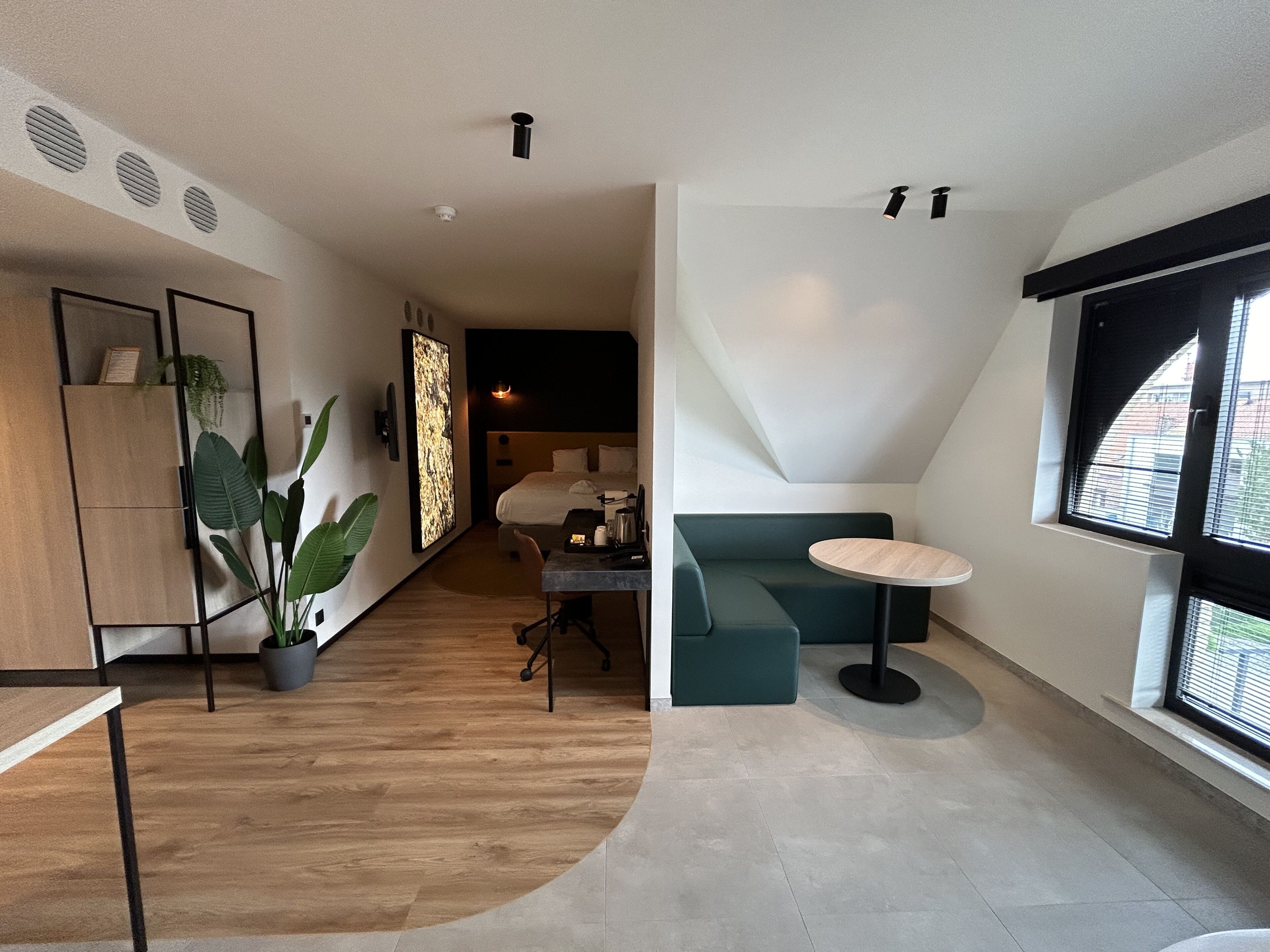 Habitació d'hotel moderna amb sòl de fusta, un sofà verd, una taula rodona petita i una finestra gran amb persianes.