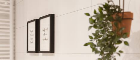 Dos letreros enmarcados sobre una pared de azulejos blancos, con una planta en maceta a la derecha.