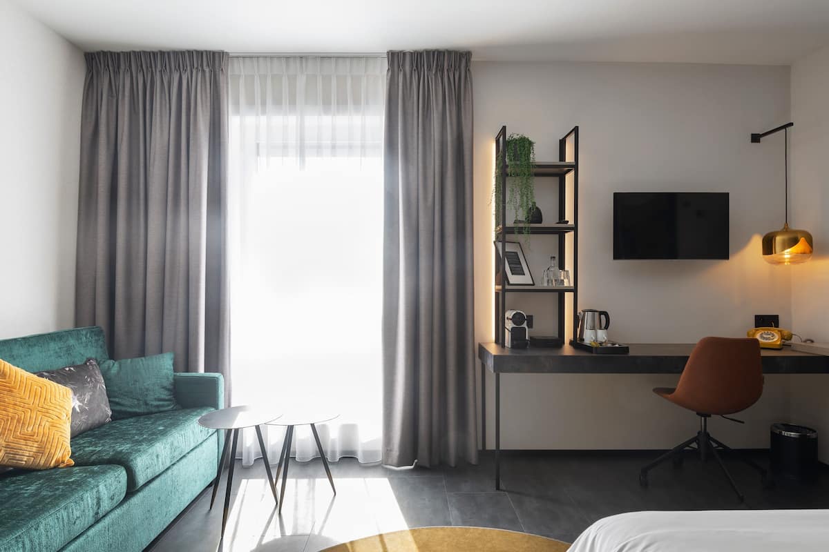 A modern hotel room with a green sofa, a small round table, a desk with a chair, a TV, and a plant on a shelf.