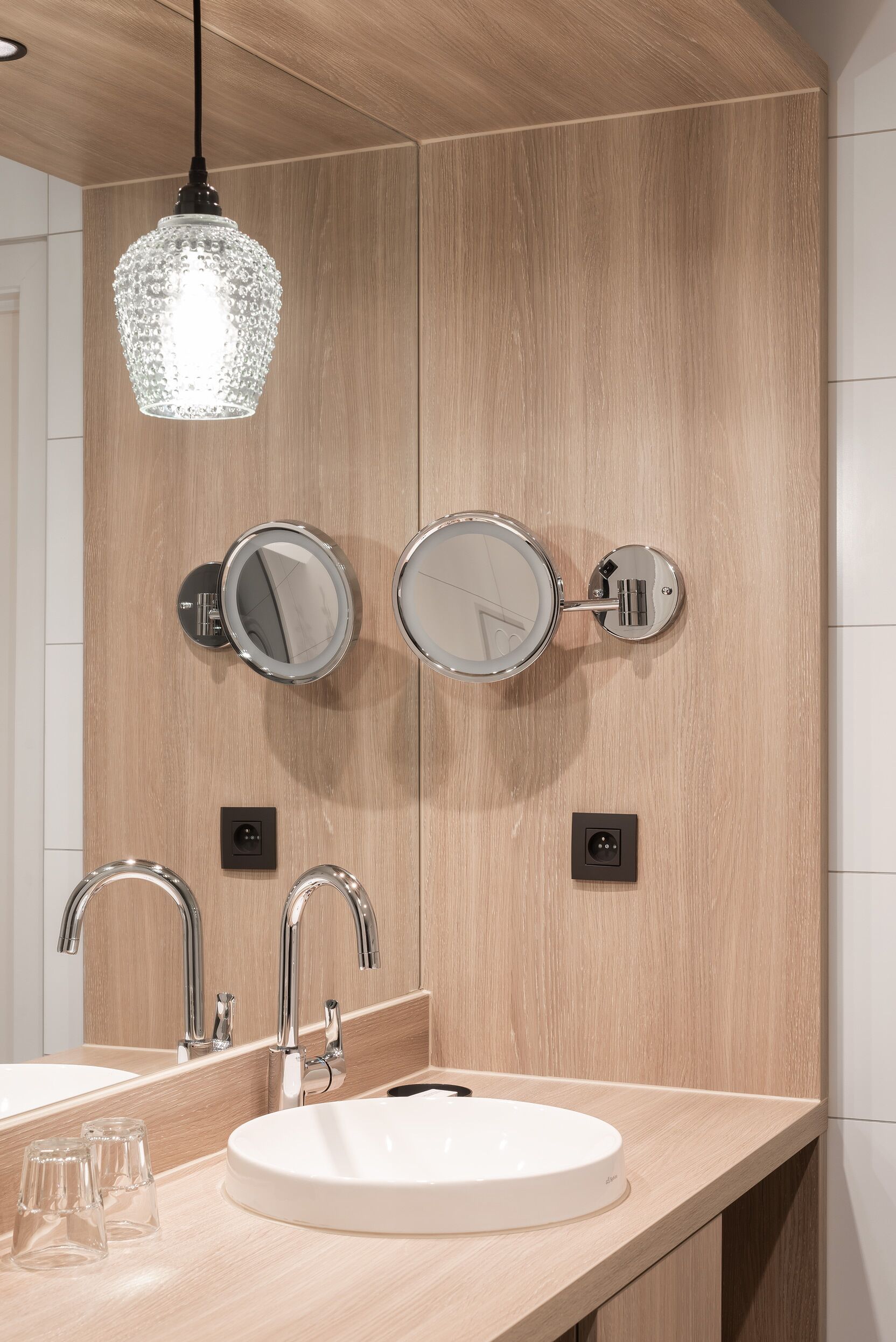 A modern bathroom with a wooden vanity, two wall-mounted mirrors, a round sink, and a pendant light.