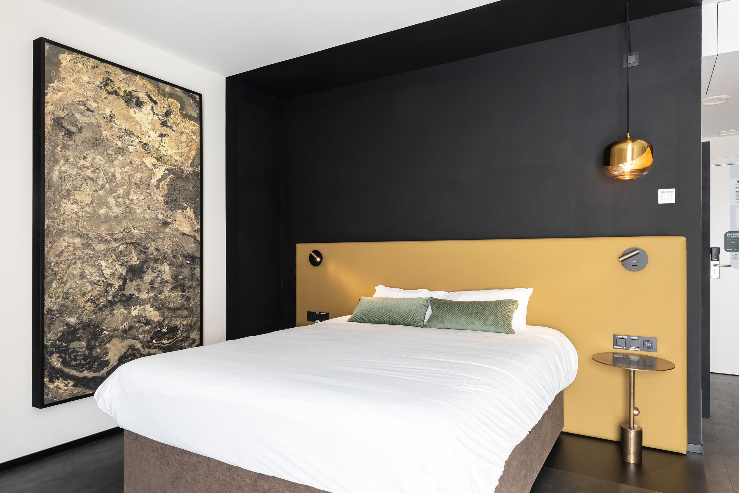 A modern hotel room with a large bed, a wall-mounted artwork, a round table with a lamp, and a control panel.