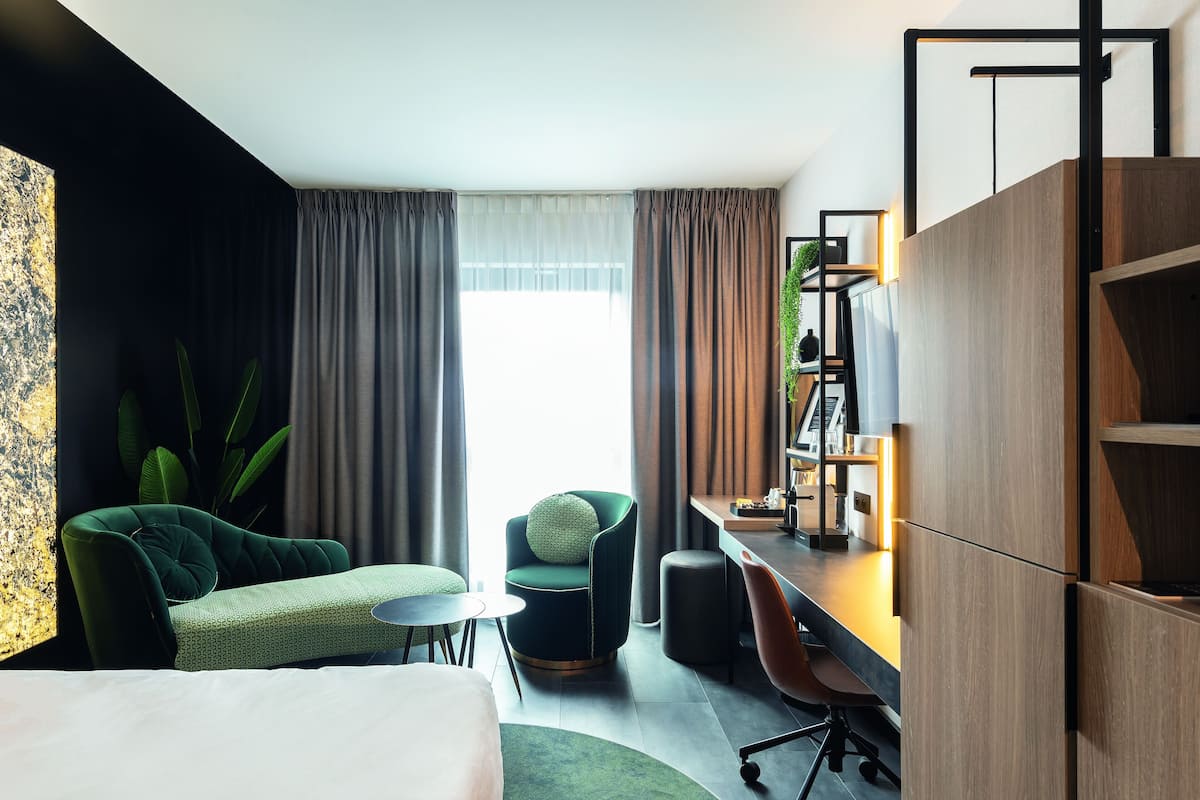 A modern hotel room with a bed, a green armchair, a desk with a computer, and a bookshelf.