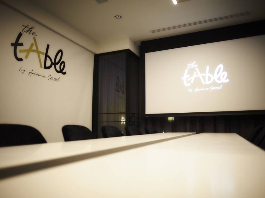A conference room with a long table and chairs, a large screen displaying 'the table by Arane Hotel' logo, and a dark-colored wall.