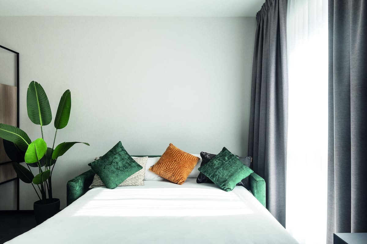 A neatly made bed with a white sheet, flanked by green and brown pillows, in a room with a large potted plant and grey curtains.
