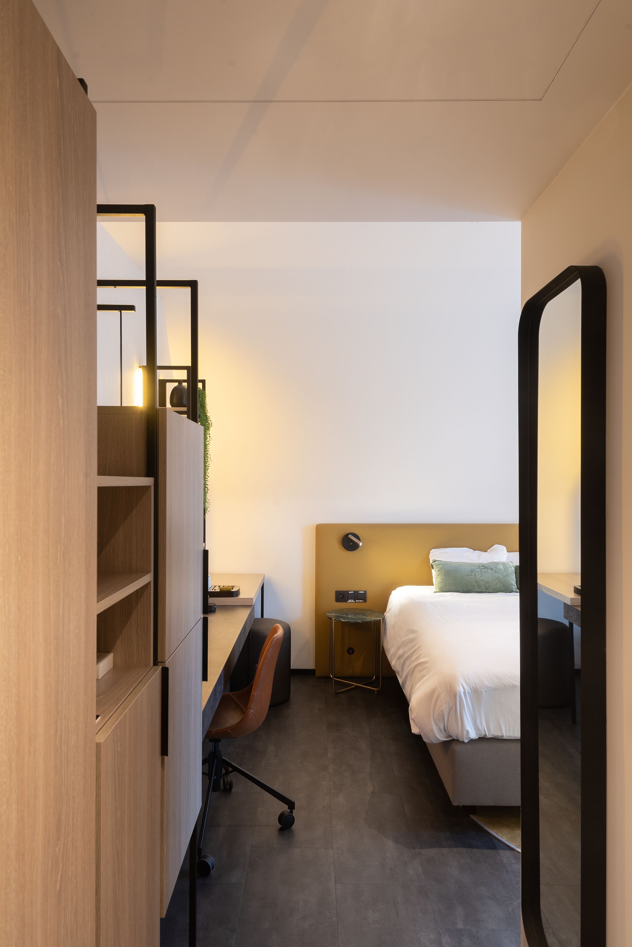 A modern hotel room with a bed, a desk, a chair, a nightstand, and a mirror.