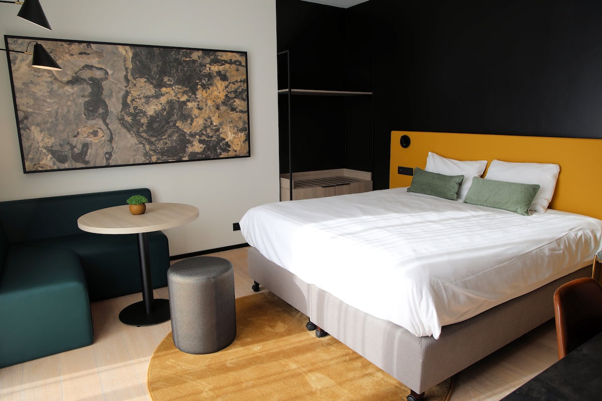 A modern hotel room with a large bed, a small round table with a plant, a sofa, and a trash bin.