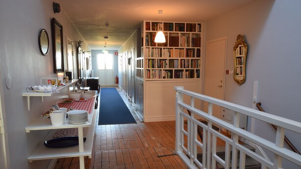 A hallway with a wooden floor, a white railing, a bookshelf, and a table with dishes and a red and white checkered tablecloth. - Bläsinge Gård Kullabygden - Hostel (Jonstorp)