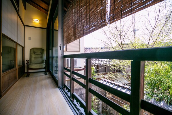 A room with a sliding door leading to a balcony, a chair, and a view of a garden and buildings outside.