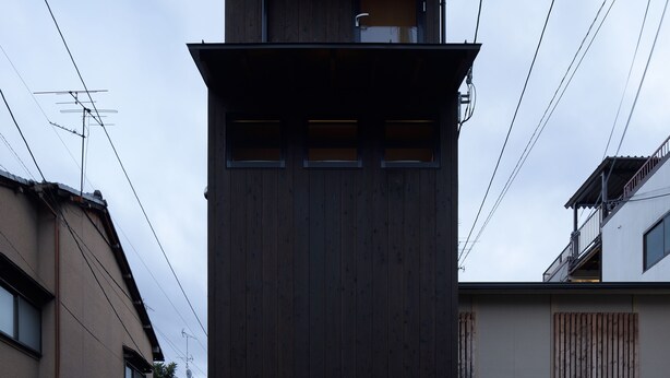 A modern two-story building with a dark wooden facade, a visible entrance with a lighted awning, and a small outdoor area with a bench and a trash bin. - KYOTO yadomachi Kamishichiken (Kyoto)