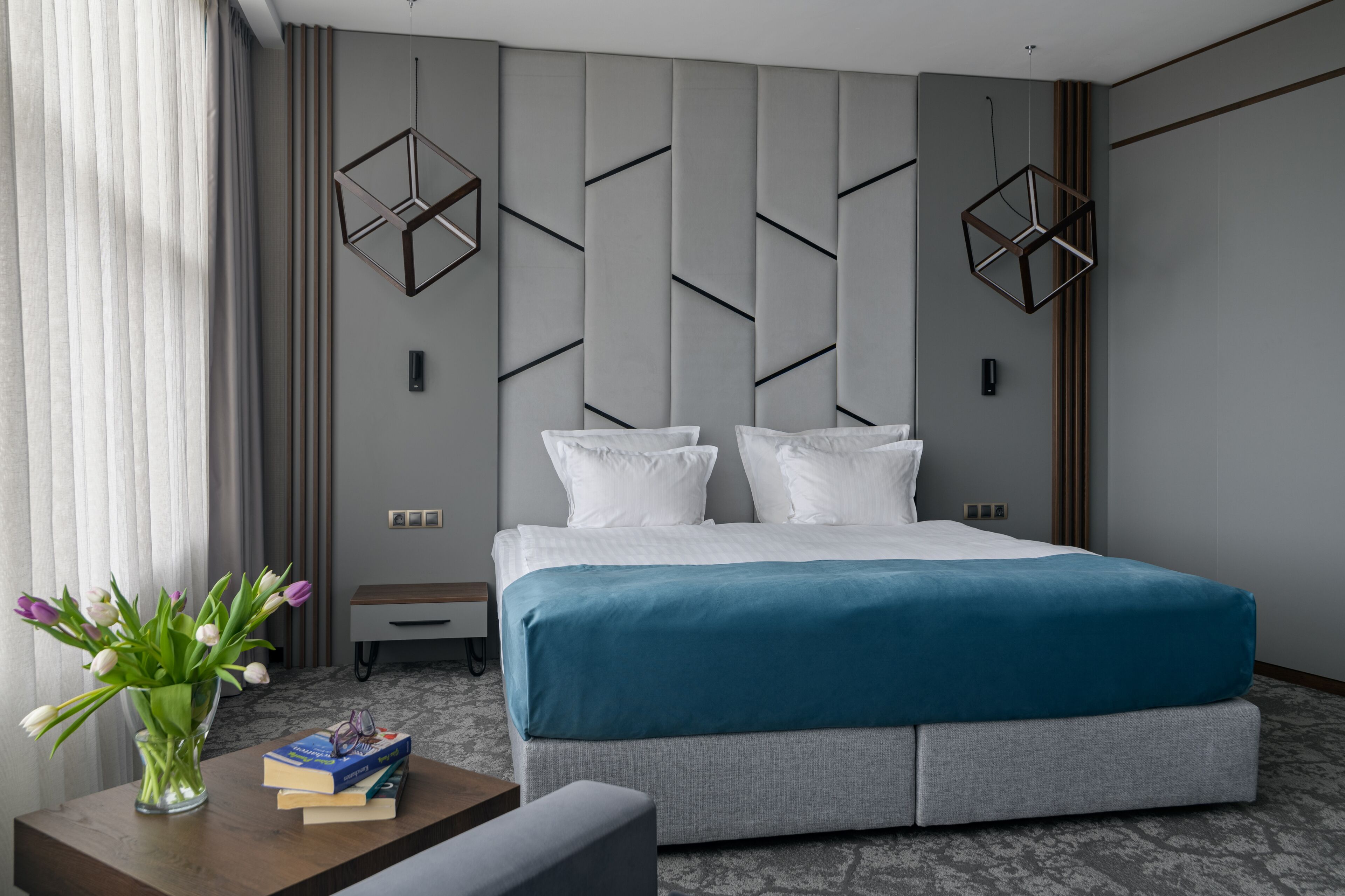 A modern hotel room with a large bed, a bedside table with books and a vase of flowers, and geometric wall panels.