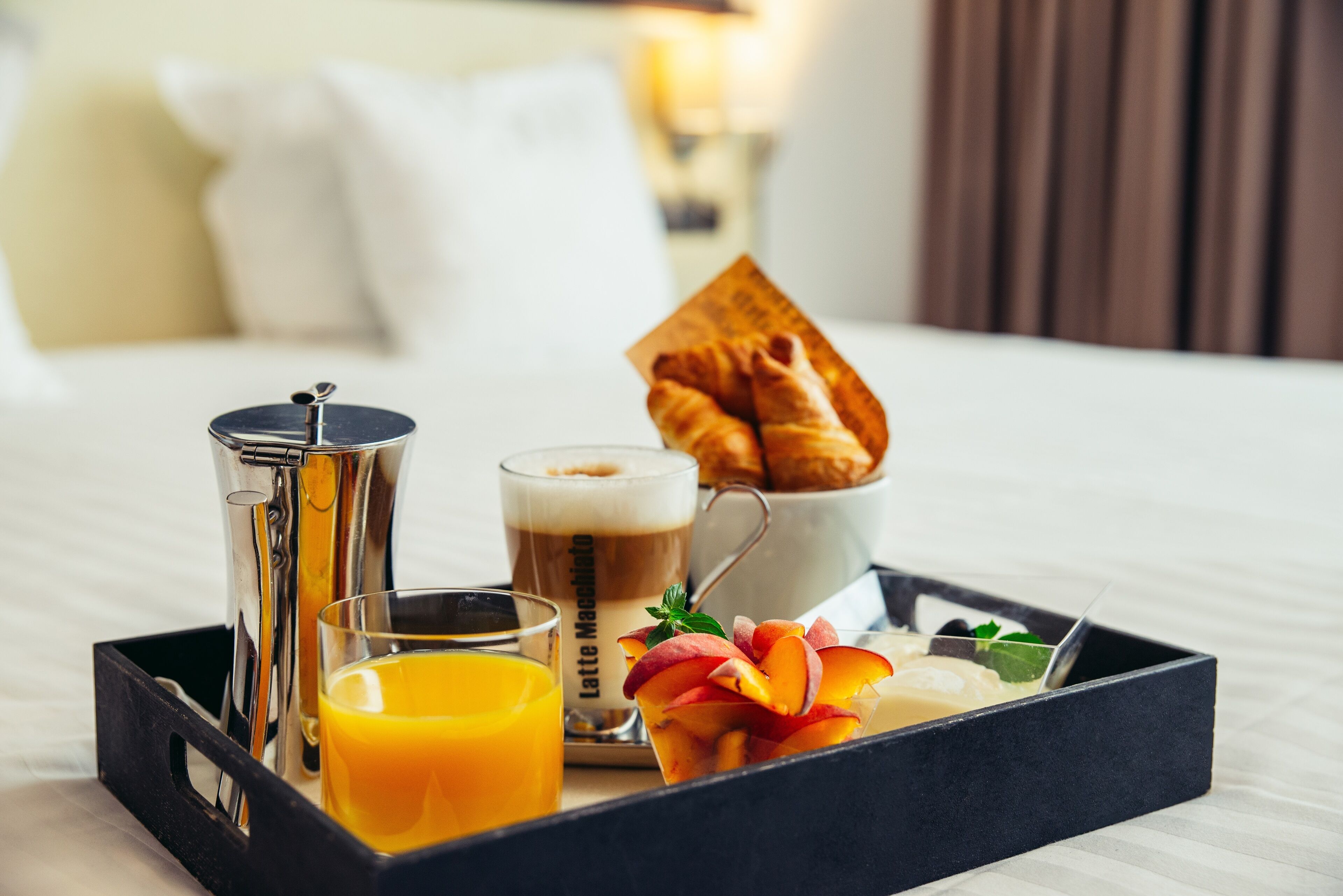 Un plateau de petit-déjeuner avec une cafetière, un verre de jus d’orange, un café au lait et un croissant, le tout posé sur un lit.