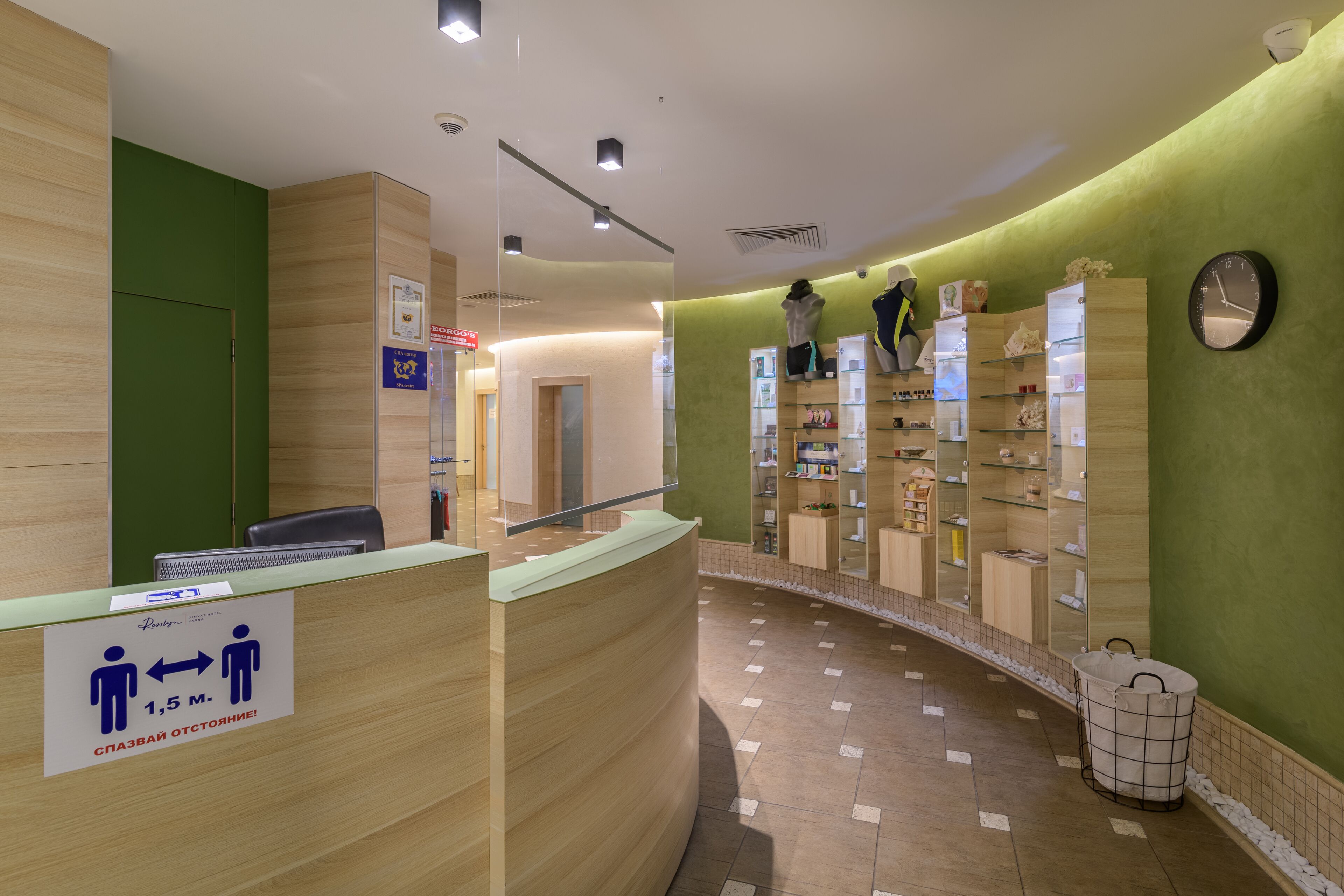 A reception area with a curved counter, a green wall, and a tiled floor. There are mannequins dressed in business attire on the wall, and a clock is visible.