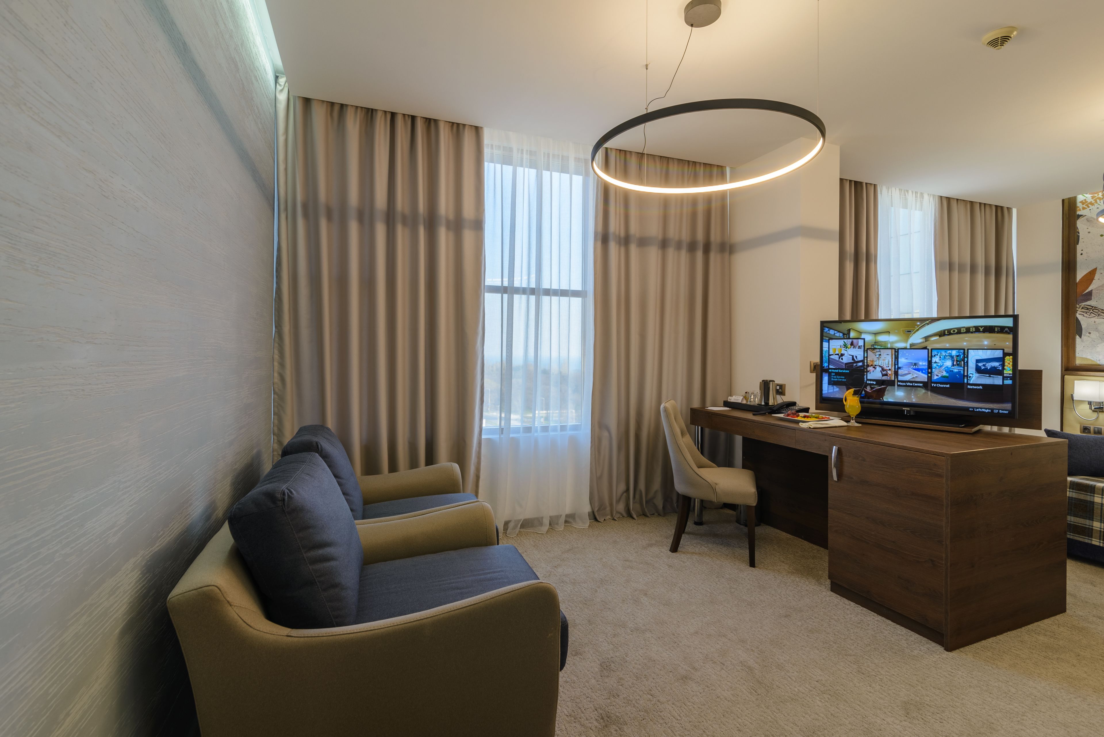 A hotel room with a seating area, a desk, a television, and a large window with curtains.