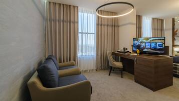 A hotel room with a seating area, a desk, a television, and a large window with curtains.