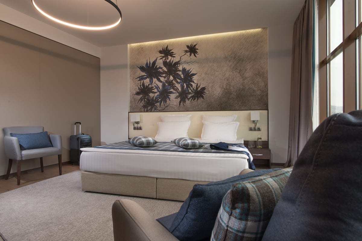 A modern hotel room with a large bed, a sofa, a chair, and a wall-mounted artwork.