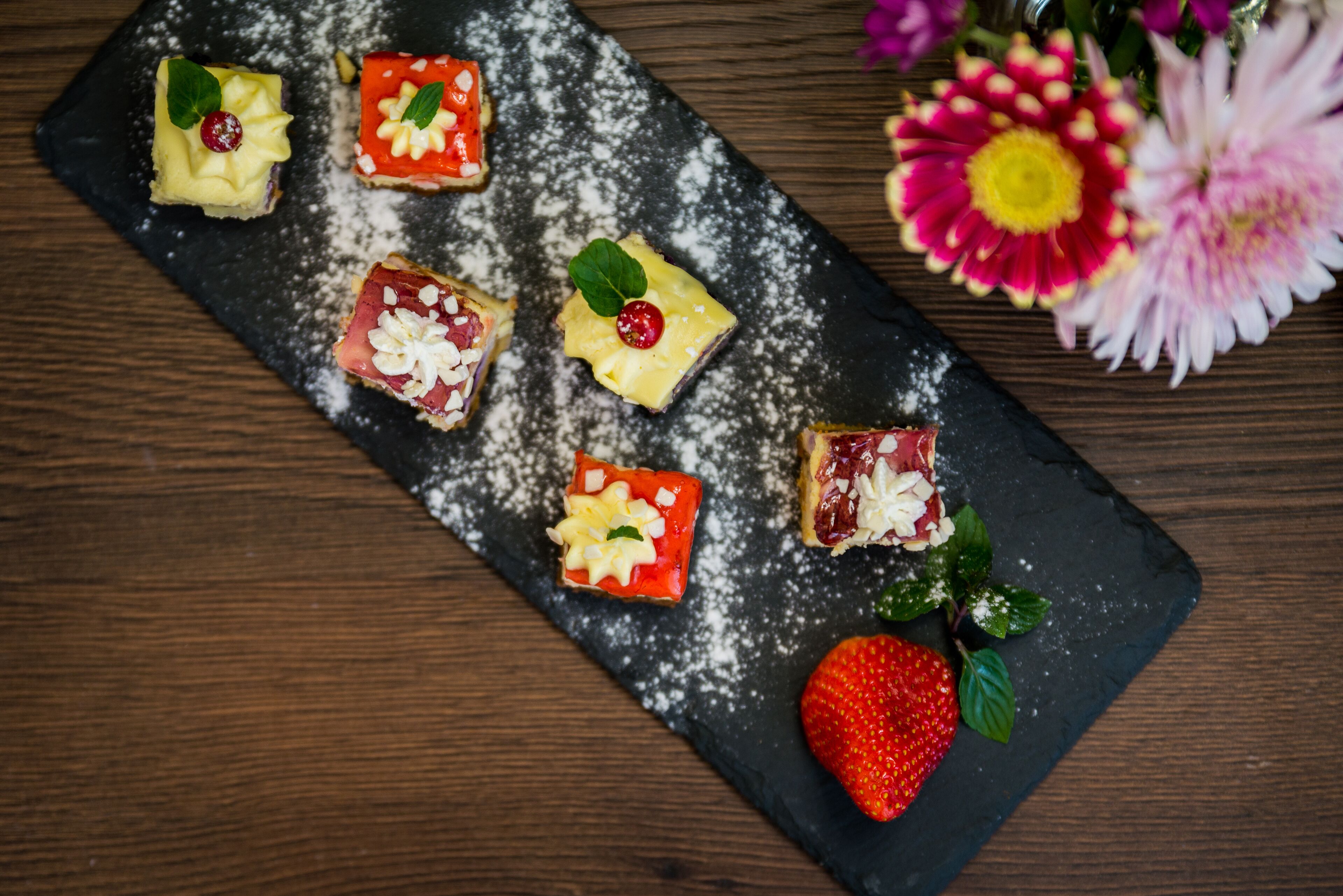 A slate serving board with six square desserts, each topped with a cherry and mint leaves, and a whole strawberry with leaves.