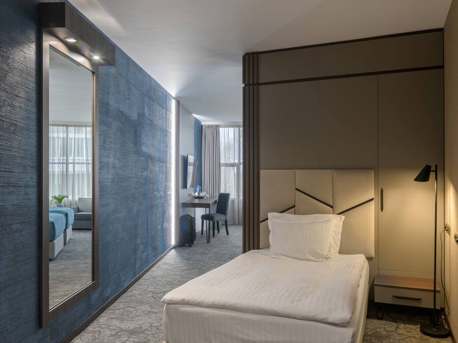 A modern hotel room with a large bed, a desk, a chair, a wardrobe, and a mirror.