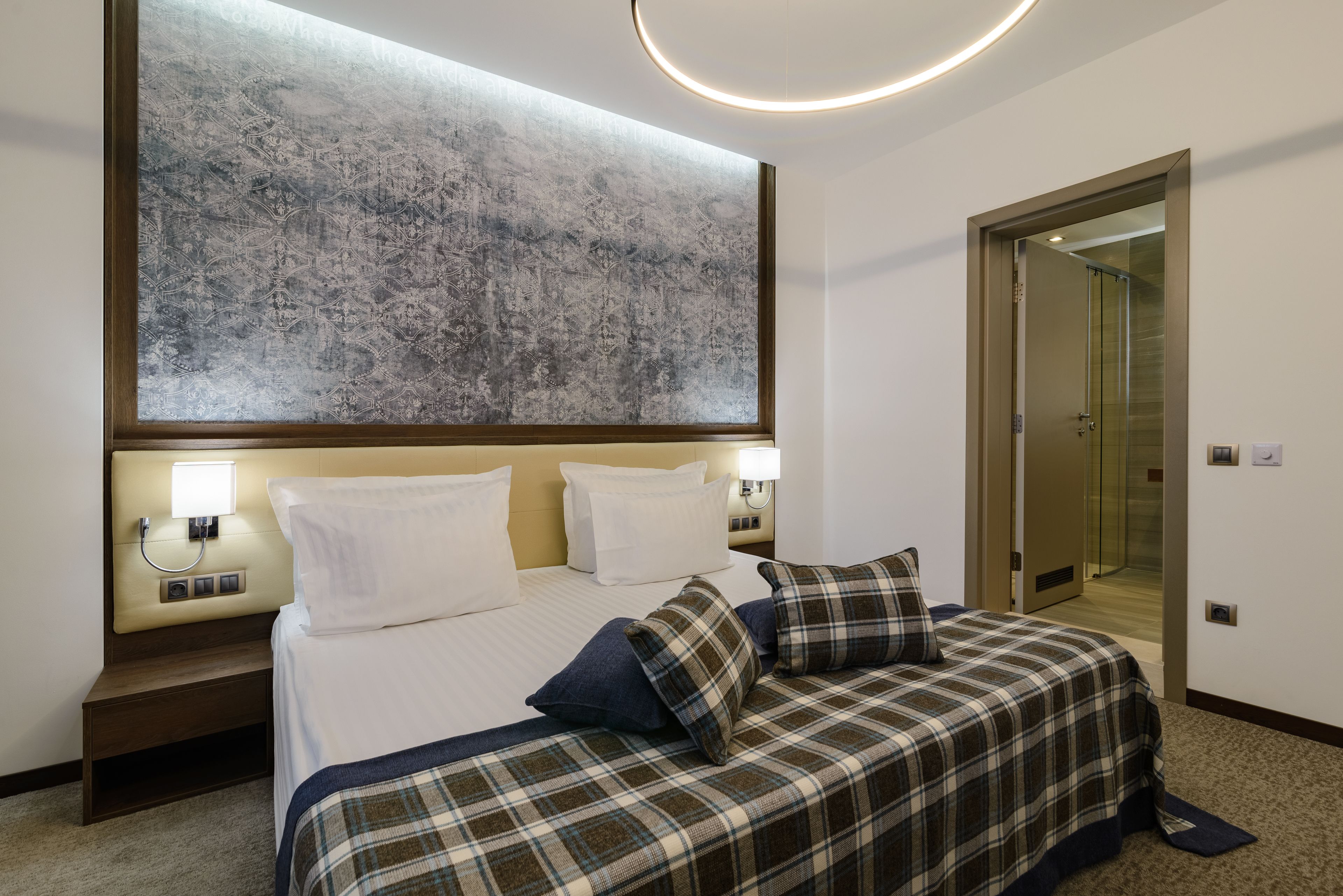 A hotel room with a large bed, a headboard with a light, a wall-mounted artwork, a bedside table with a lamp, and a door leading to a bathroom.