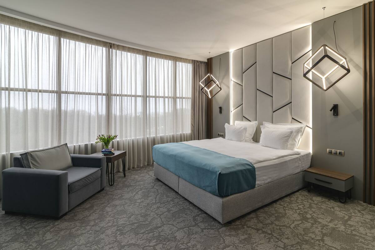 A modern hotel room with a large bed, a sofa, a small table, and a potted plant.