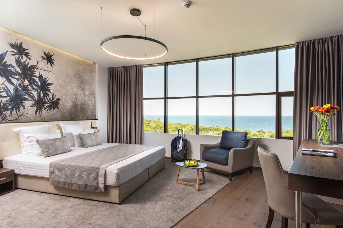 A modern hotel room with a large bed, a desk, a chair, and a view of the ocean.