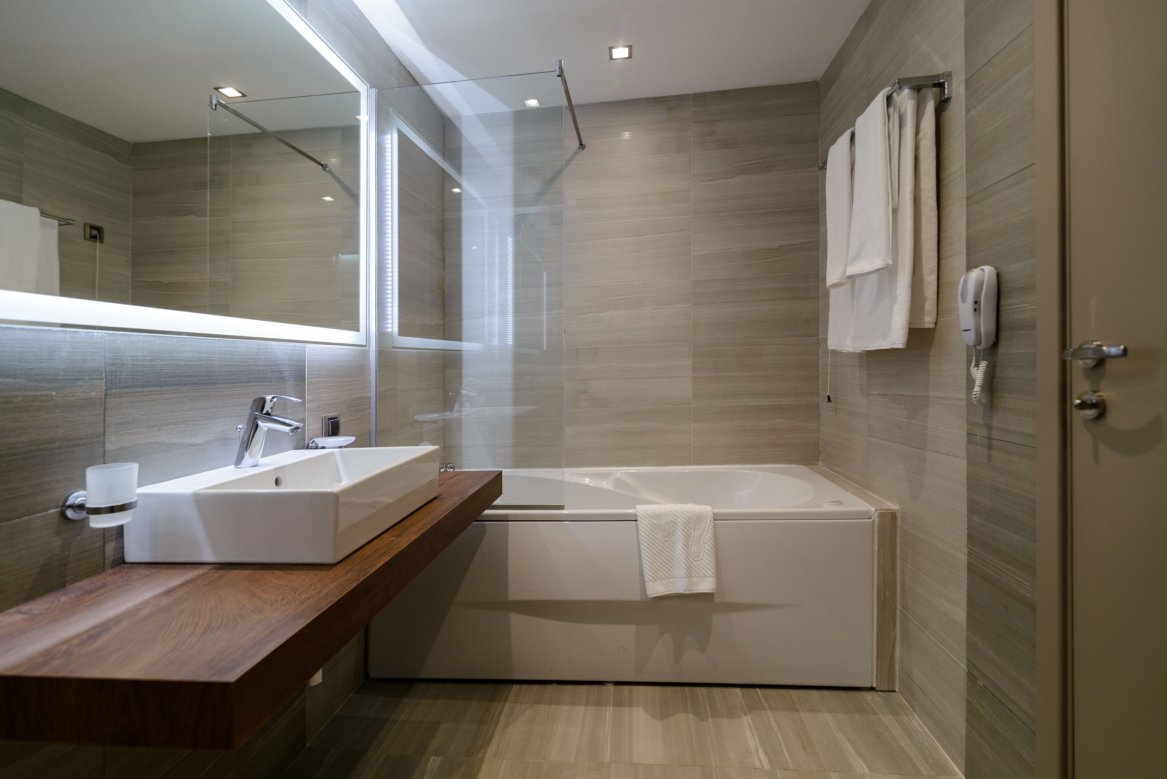 A modern bathroom with a large mirror, a wooden vanity, a white bathtub, and a glass-enclosed shower area.
